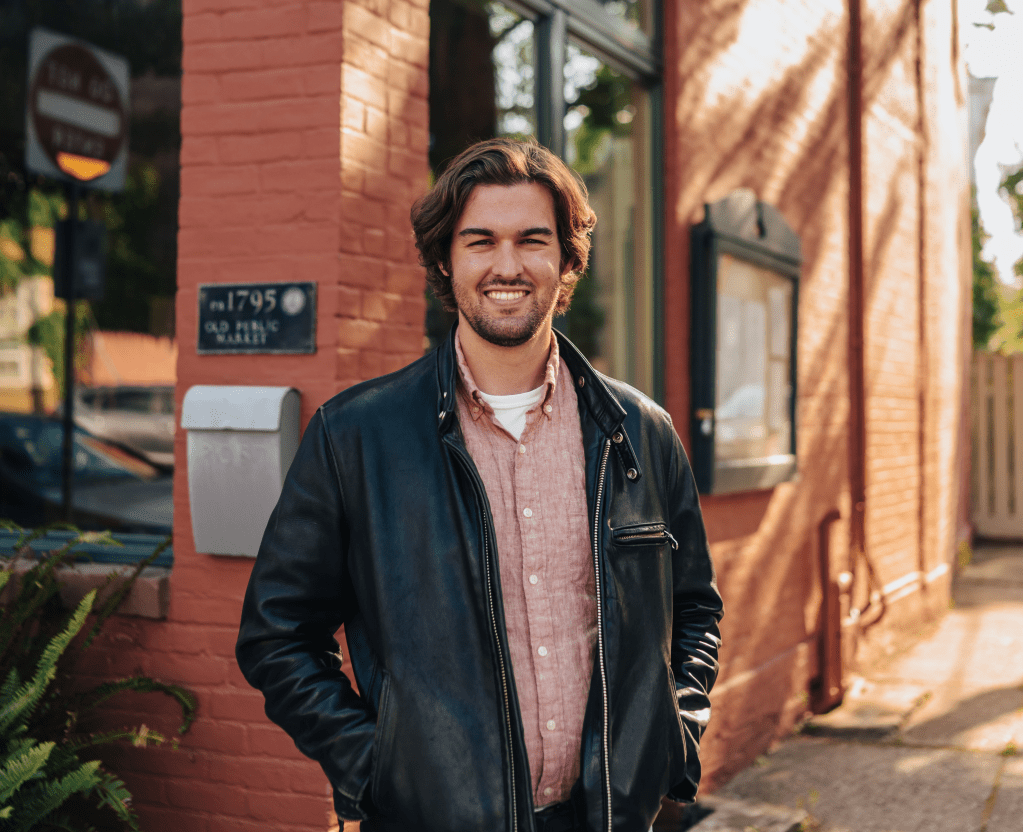 Medium-distance photo of a man in a dark jacket, smiling, on a sunny day.