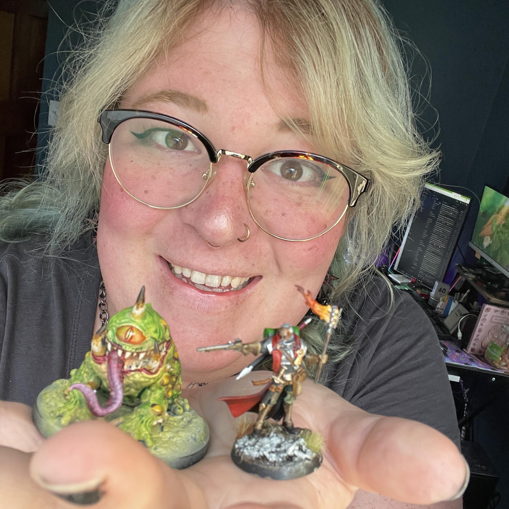 Close up of a white-presenting woman with blonde hair and glasses holding two painted minis to the camera.