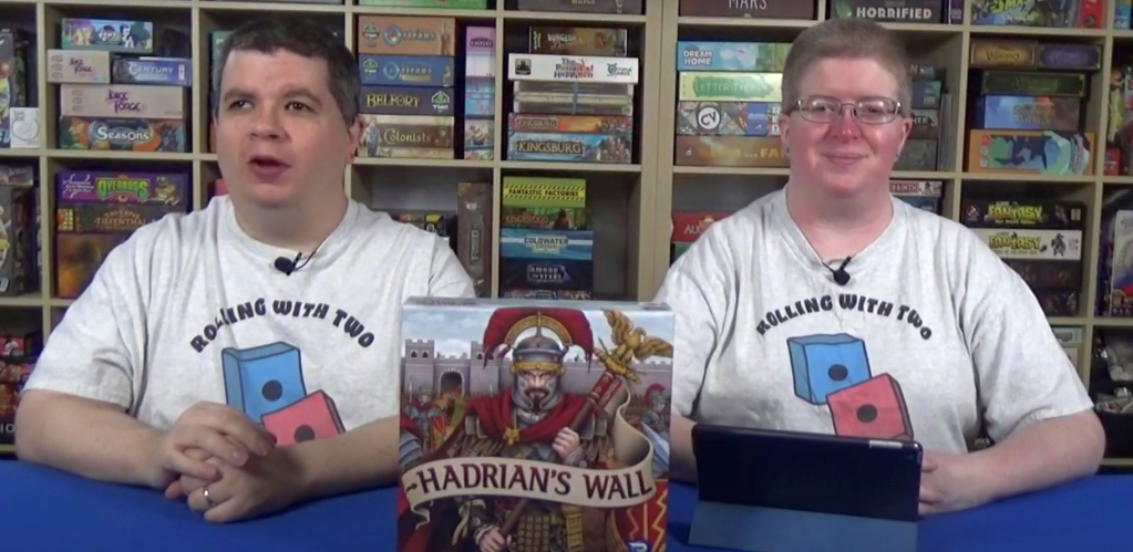 Image of two people sitting at a table, looking at the camera. The Hadrian's Wall game box is in the foreground between then. Each person (male-presenting on left; female-presenting on right) are wearing white T-shirts with the words "Rolling With Two" and cartoon-art of two dice printed on the shirts.